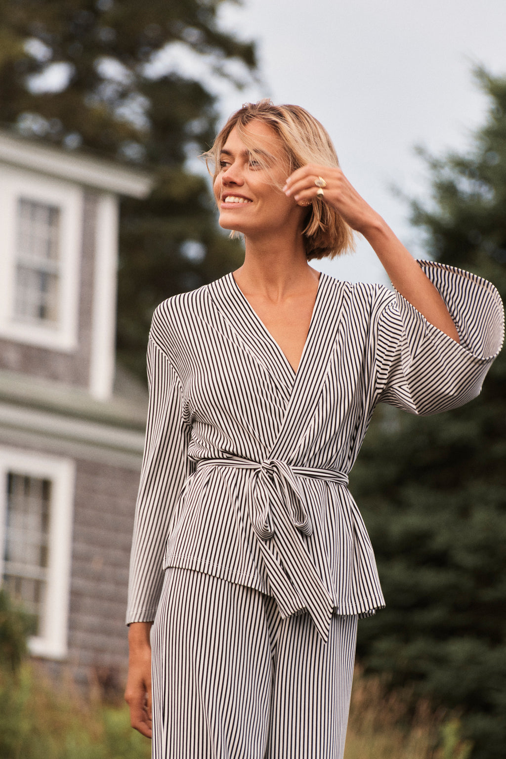 A woman wears the DreamModal Kimono Pajama Set in Black Pencil Stripe outdoors, smiling, with a house featuring white trim and greenery in the background.