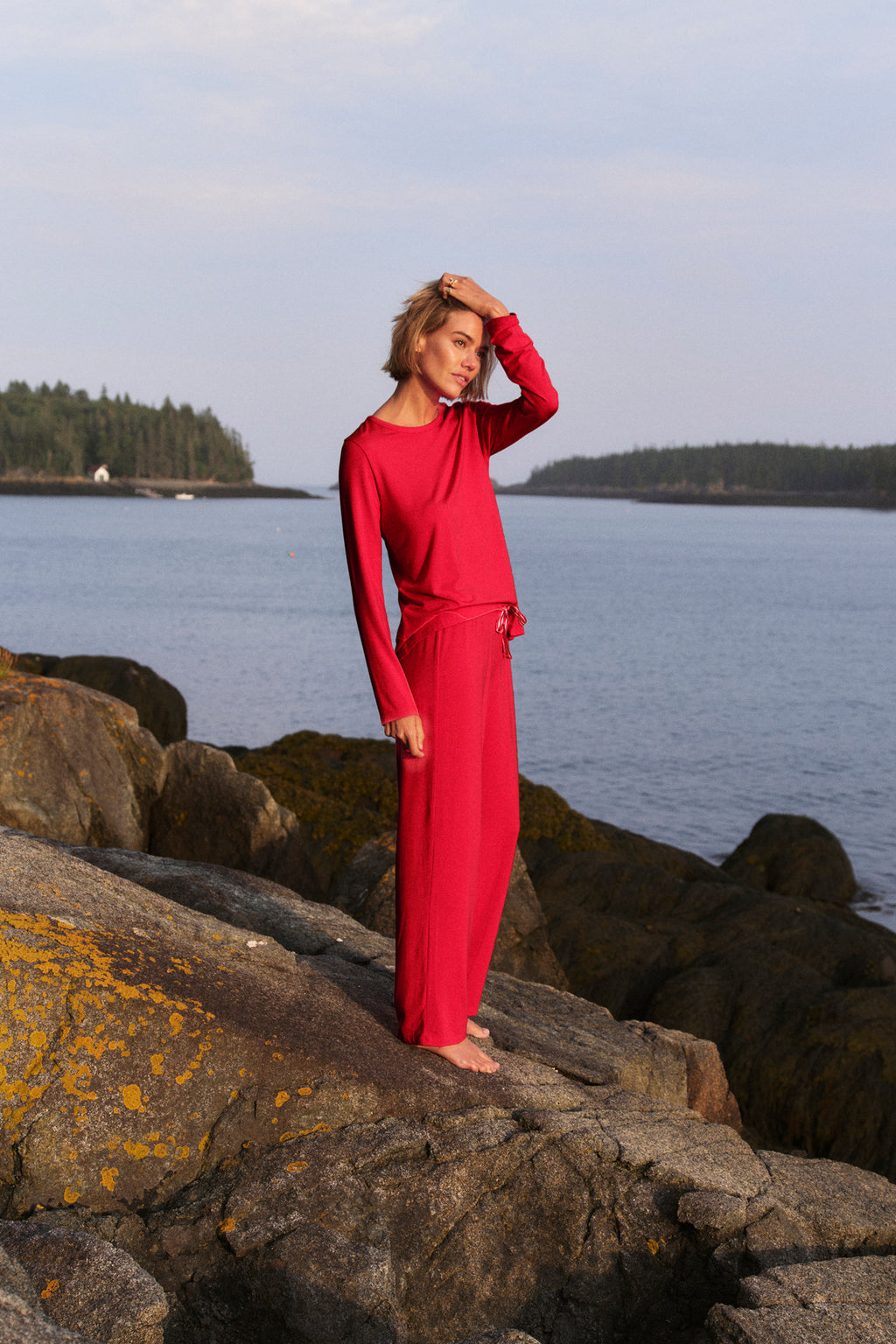 Wearing DreamModal Ribbon Long-Long Set in Ski Patrol, a person in red drawstring pajama pants stands by rocky water with trees and a small building across, under a partly cloudy sky, one hand touching their head.