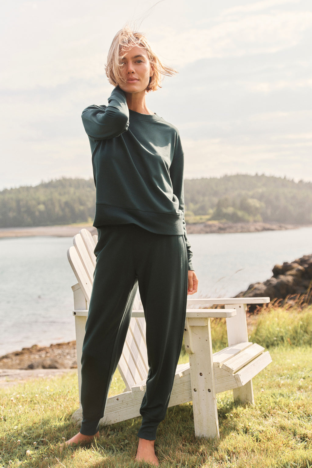 A person stands on grass by a wooden chair near water, wearing the Relax Jogger Set in Conifer, with gentle sunlight and trees in the background.