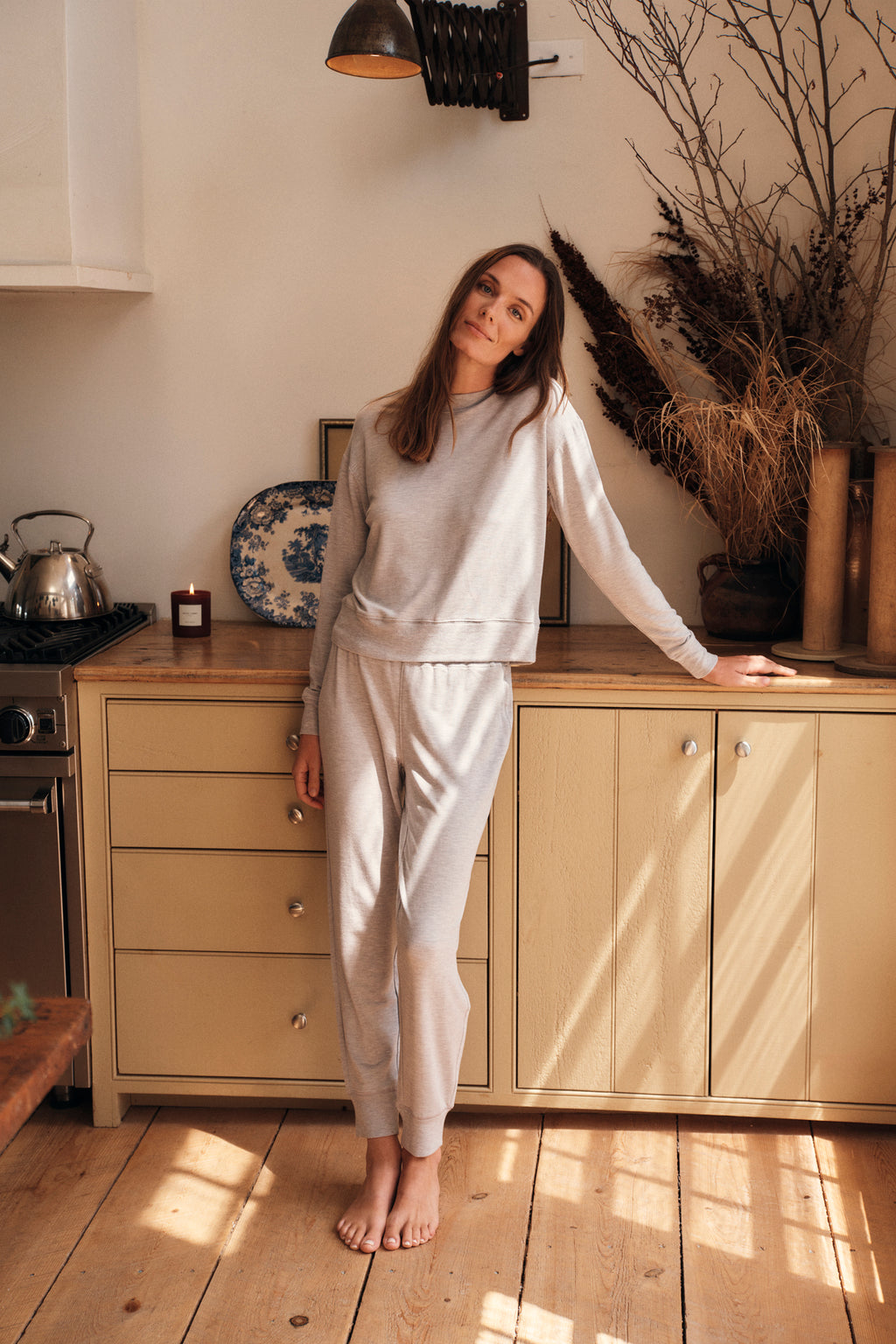 A woman wearing the Relax Jogger Set in Heather Gray stands in a cozy kitchen, leaning against a yellow cabinet. Dried plants and pottery decorate the space as sunlight streams through, creating a warm atmosphere.
