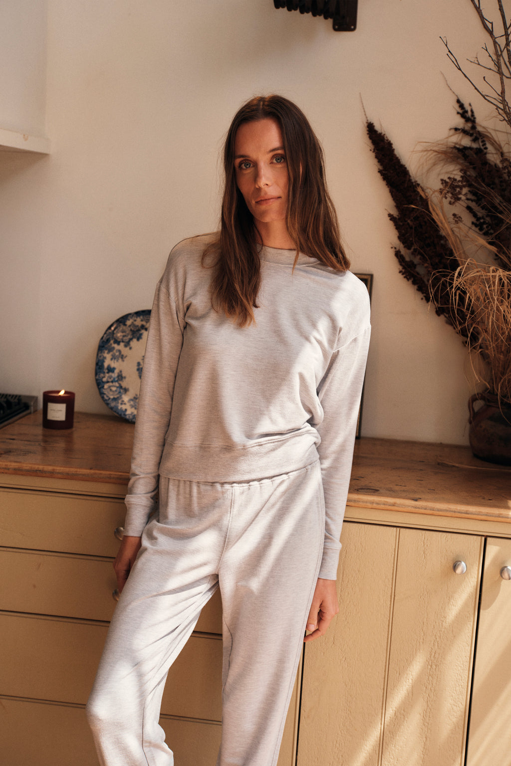 A woman wearing the Relax Jogger Set in Heather Gray stands by a wooden cabinet with a decorative plate, candle, and dried plants behind her, looking at the camera with a relaxed expression.