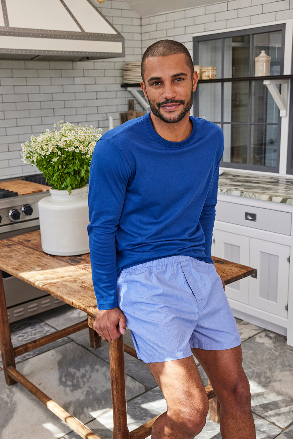 A bearded man in a blue long-sleeve shirt and Men’s Poplin Boxer Bundle in Blue sits on a wooden kitchen island in a bright kitchen with white cabinets and a vase of white flowers.
