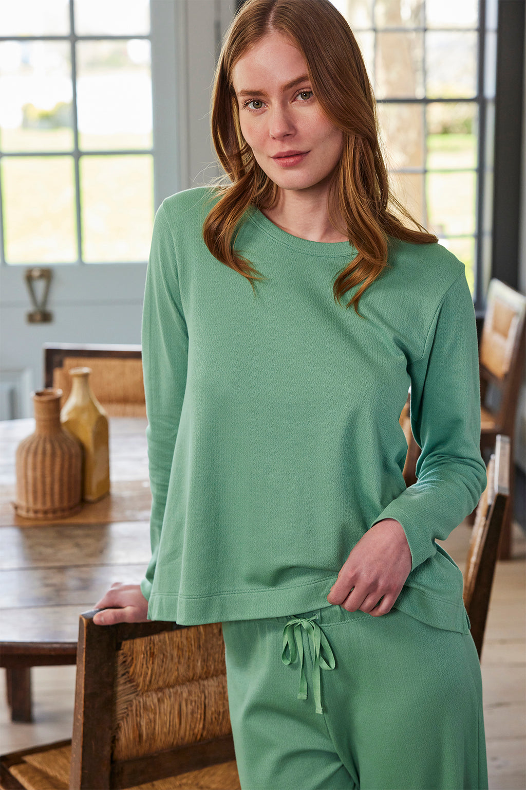 A woman in the Pointelle Pants Bundle in Boxwood stands indoors by a wooden dining table, with bottles and chairs visible in the background.