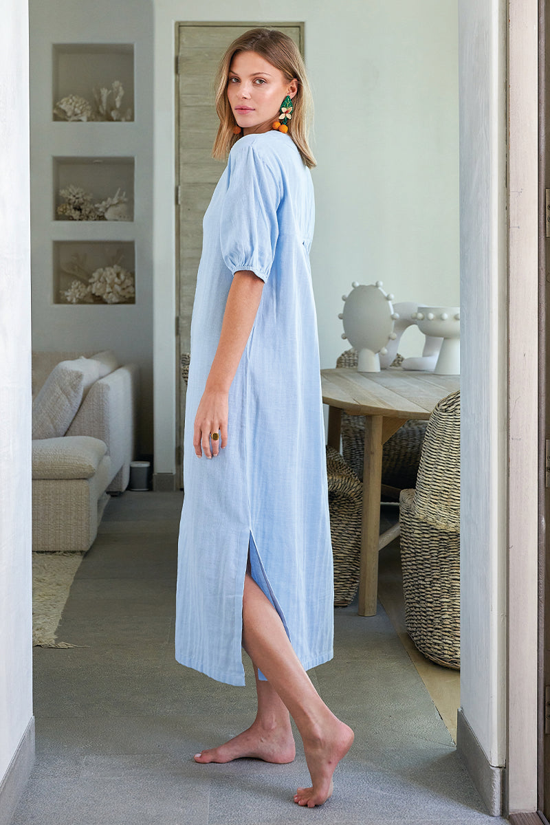 A woman wears the Gauze Caftan in Morning Blue indoors. This loose-fitting, 100% cotton midi dress features short puff sleeves, a side slit, and offers soft comfort. She looks over her shoulder in a bright, modern room.