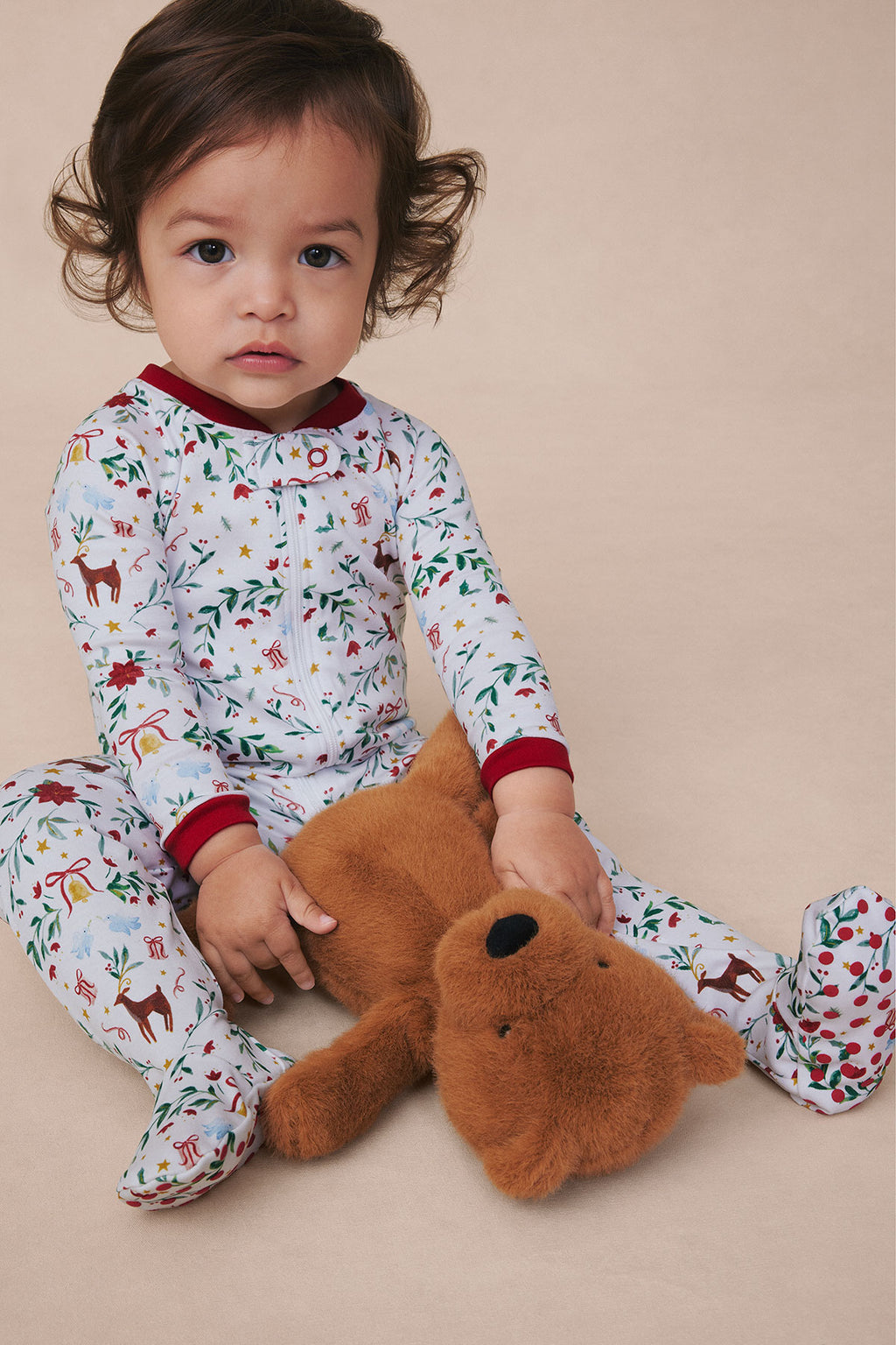 Toddler wearing white pajamas with holiday-themed print, red trim, and feet, holding a brown stuffed animal.