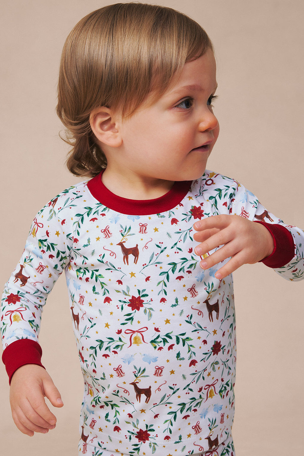 Toddler wearing long-sleeve pajama top with red trim, featuring holiday-themed print of reindeer, flowers, gifts, and stars o