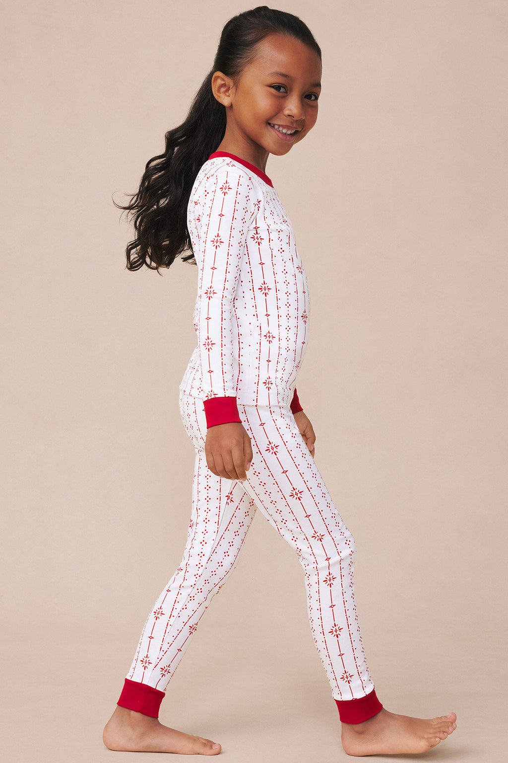 Child wearing white long-sleeve pajama top and matching pants with red geometric stripe and snowflake pattern, red cuffs at w