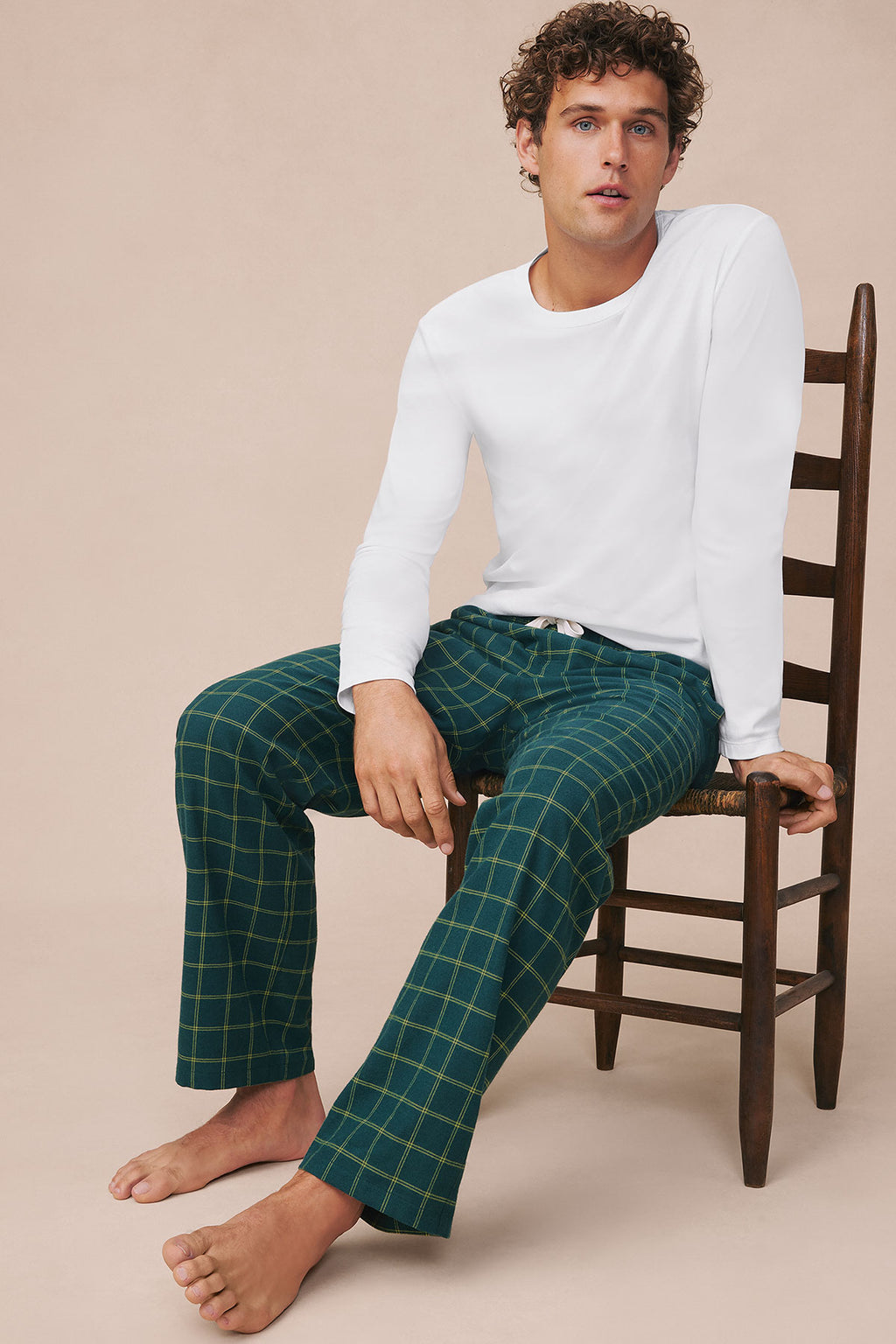 Dark teal windowpane flannel pajama pants with yellow grid pattern worn by man sitting on wooden chair.