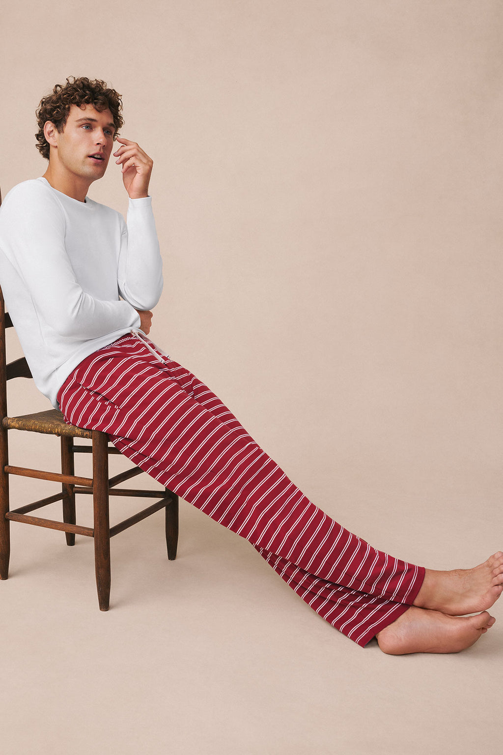 Man wearing white long-sleeve top with red and white striped long pants seated on wooden chair.