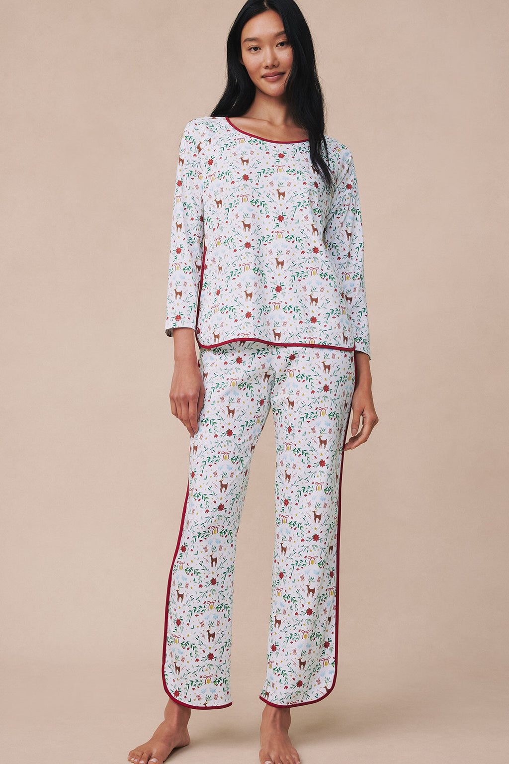 Woman wearing a long-sleeve pajama set with festive print and burgundy piping on a cream background.