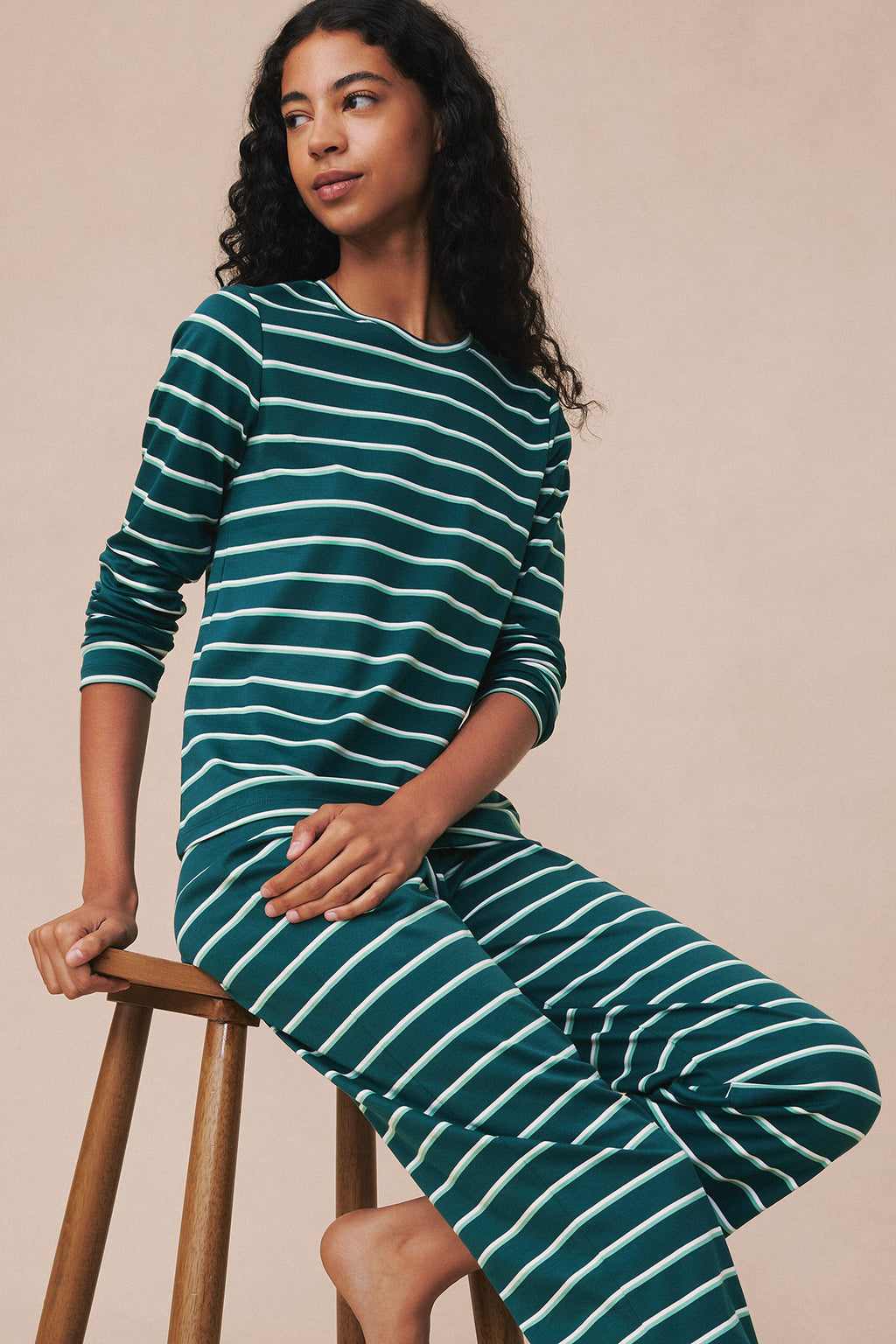 Dark teal and white striped long-sleeve crew neck top and matching pajama pants on model seated on wooden stool.