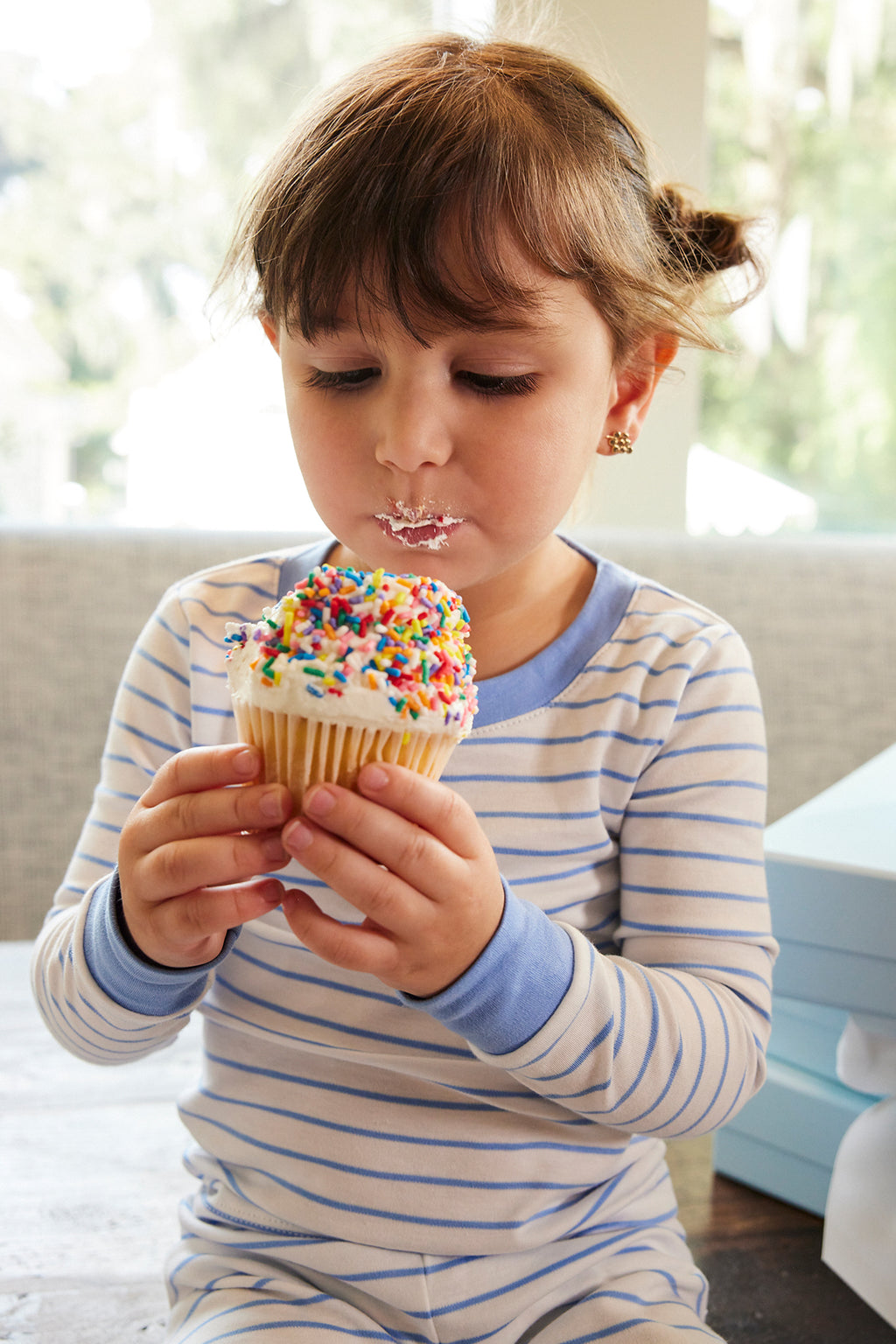 A young child wearing the Kids Long-Long Set in Baltic Blue holds a sprinkle-covered cupcake, gazing down at it with frosting on their lips.