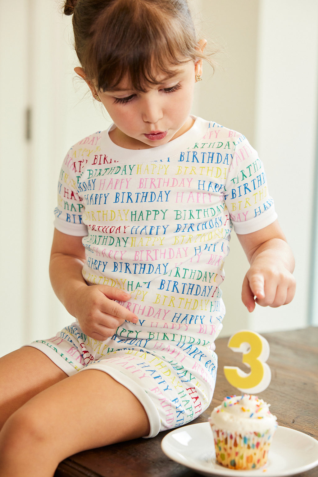 A young girl in a colorful Kids Shorts Set in Happy Birthday sits at a table, looking down at a cupcake topped with sprinkles and a yellow number 3 candle.