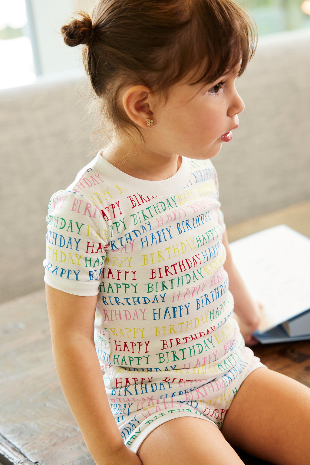 A young girl sits on a bench wearing the Kids Shorts Set in Happy Birthday—a white 100% Pima cotton outfit with colorful HAPPY BIRTHDAY text—looking to the side with her mouth slightly open. A notebook is on the nearby table.