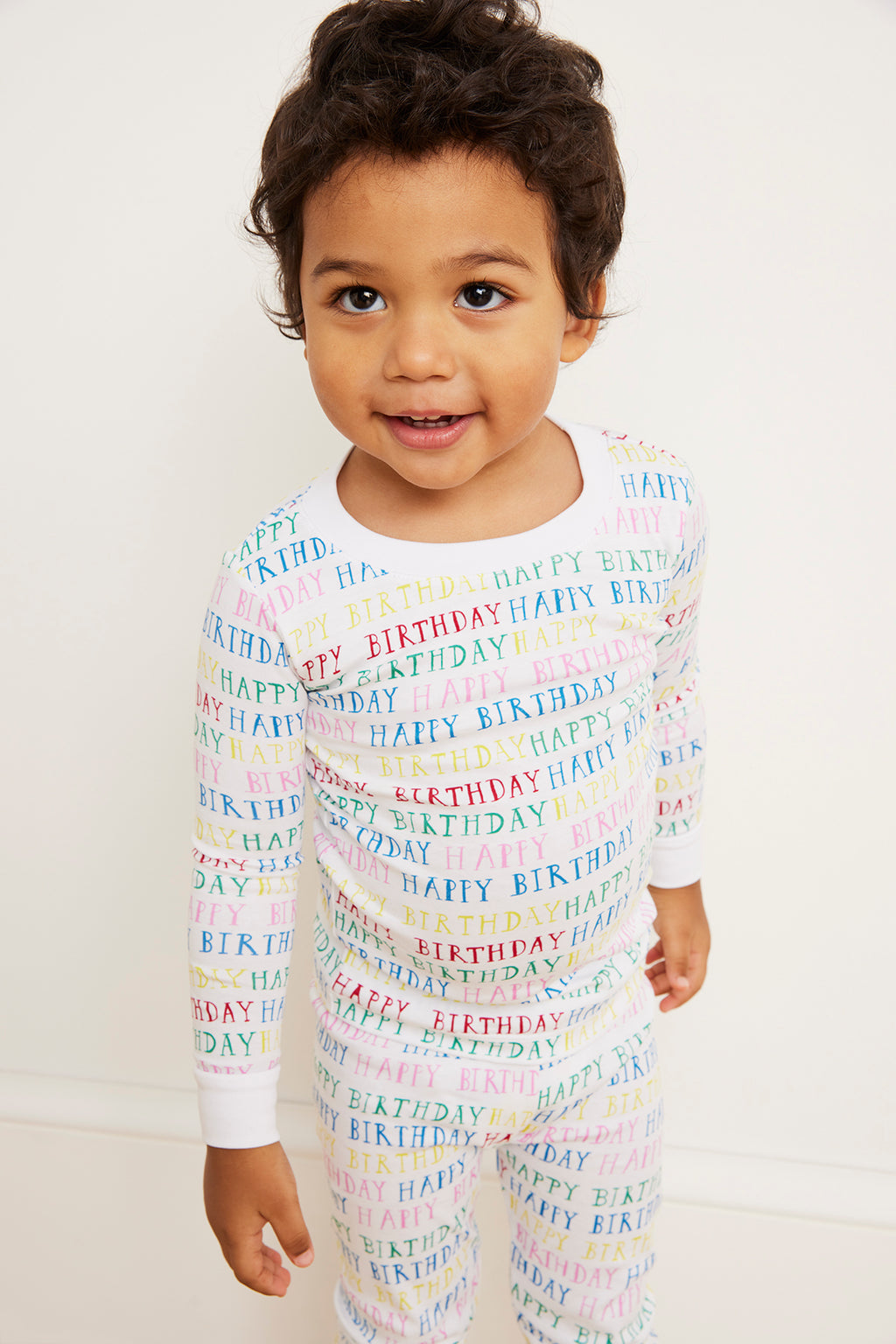 A young child smiles against a white wall, wearing the Kids Long-Long Set in Happy Birthday—white pajamas in soft Pima cotton with pastel HAPPY BIRTHDAY text and a comfy, snug fit.
