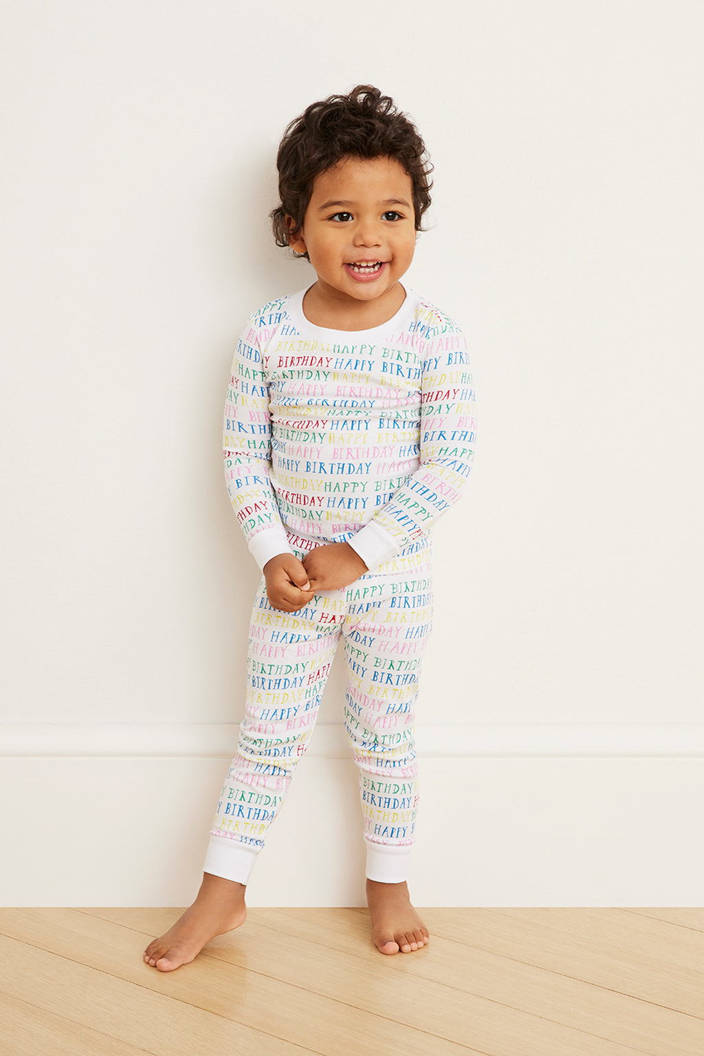 A smiling young child stands indoors, leaning against a white wall on a light wooden floor, wearing the Kids Long-Long Set in Happy Birthday—crafted from soft Pima cotton and printed with colorful HAPPY BIRTHDAY text.