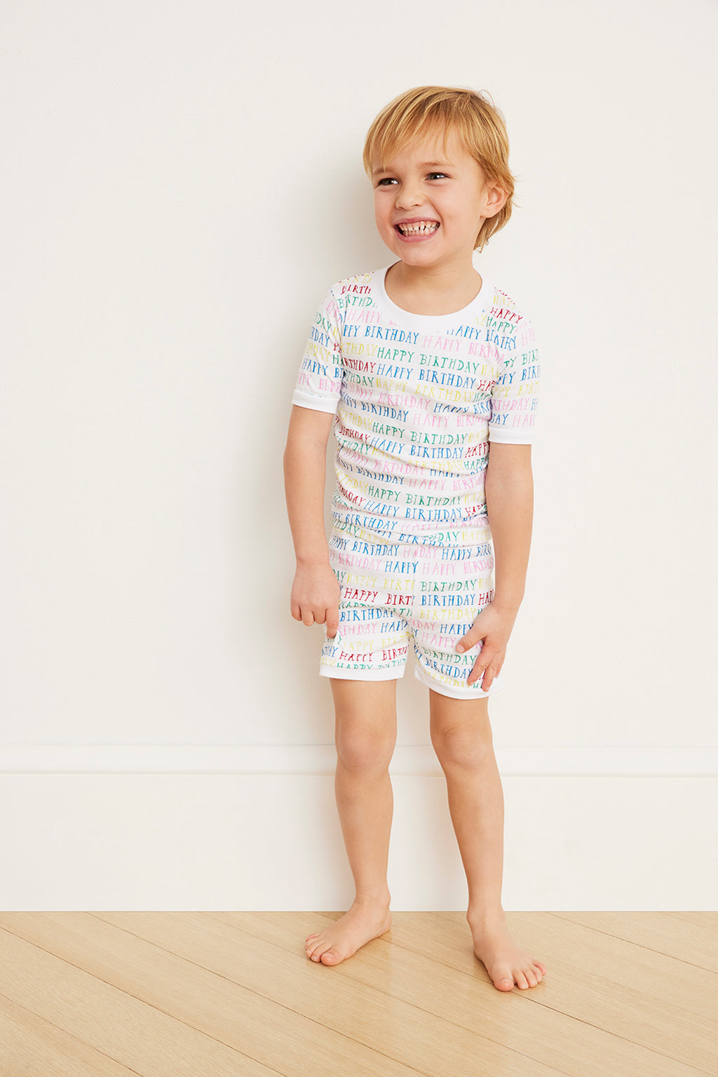 A young child smiles against a white wall, wearing the Kids Shorts Set in Happy Birthday—crafted from 100% Pima cotton with an elastic waistband and a colorful birthday print.