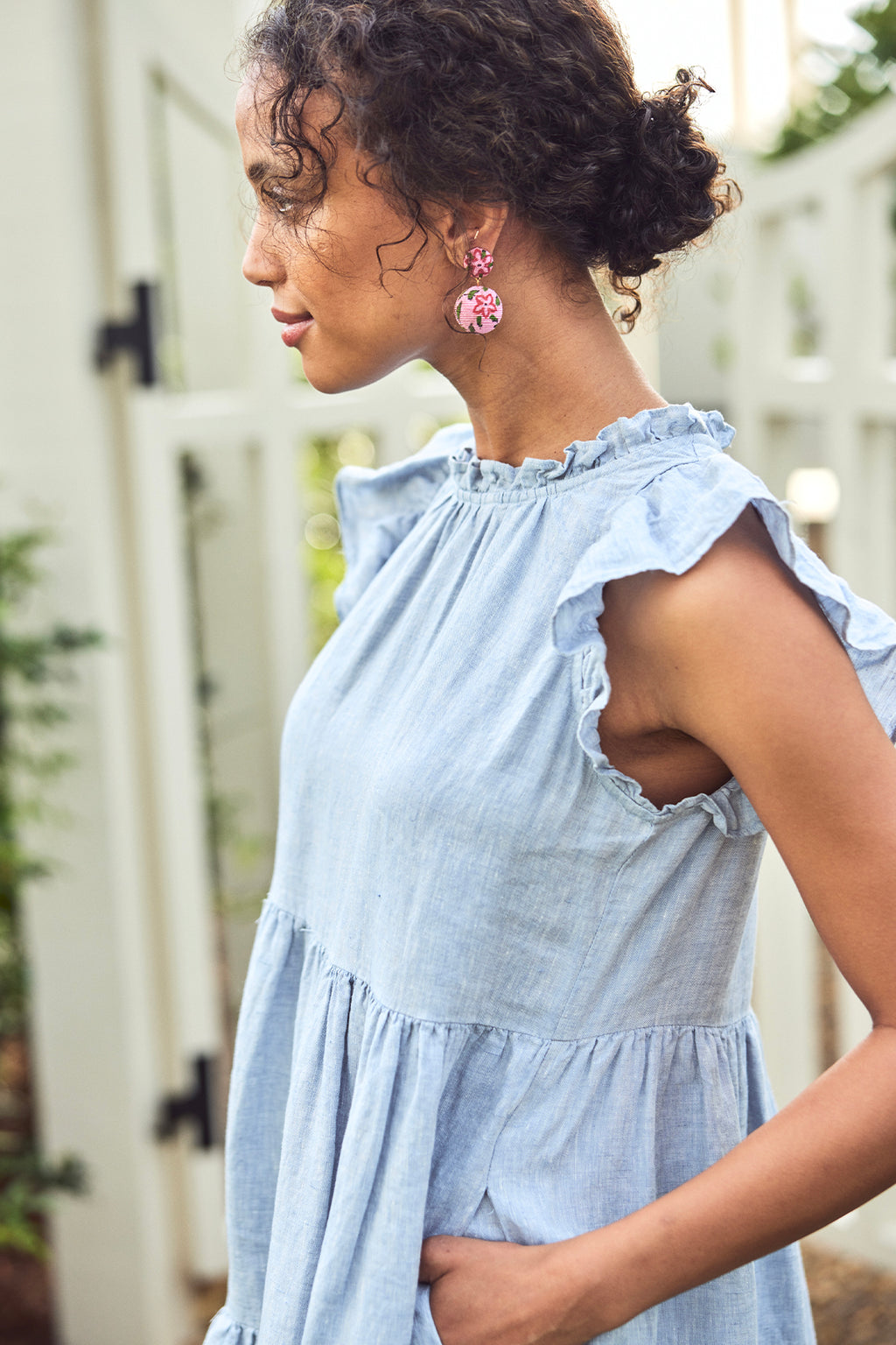 Woman stands outdoors by a white fence, viewed from the side, wearing the Helen Dress in Blue Linen with ruffled tiers and pink floral earrings.