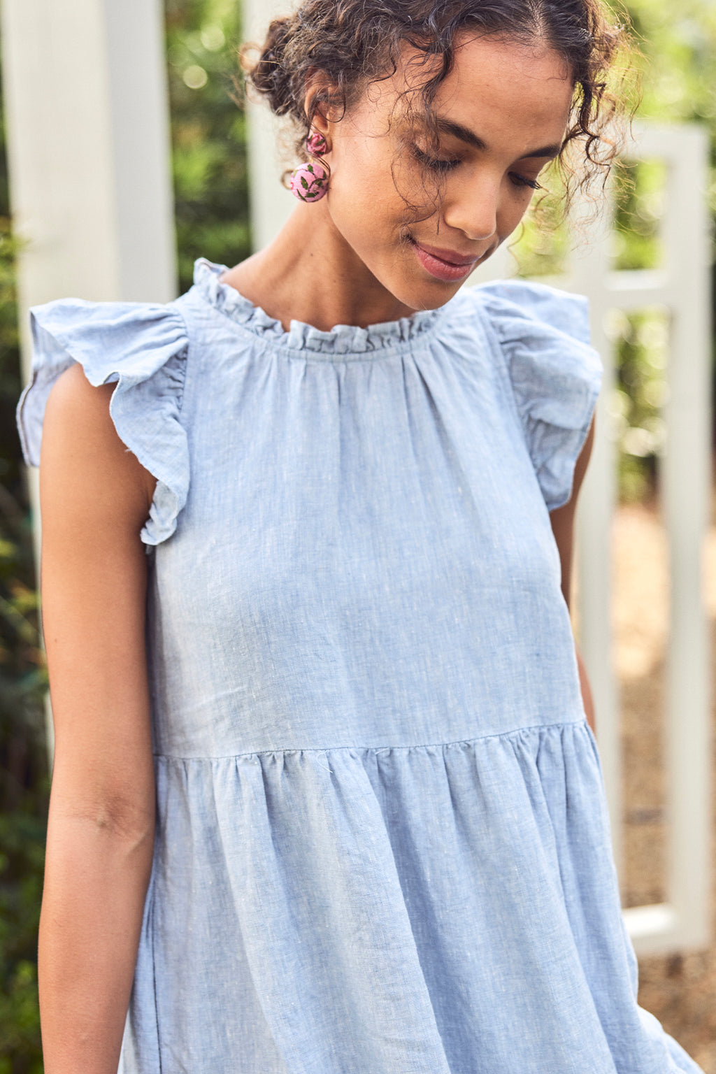 Wearing the Helen Dress in Blue Linen with ruffled sleeves and pink earrings, a woman stands outdoors by greenery and a white fence, looking down with a gentle smile.