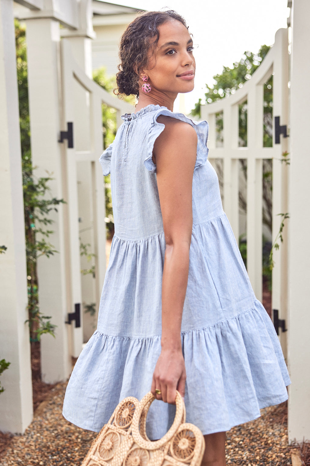 A woman in the Helen Dress in Blue Linen stands outdoors by a white wooden gate, holding a woven straw bag and wearing pink earrings, looking over her shoulder.