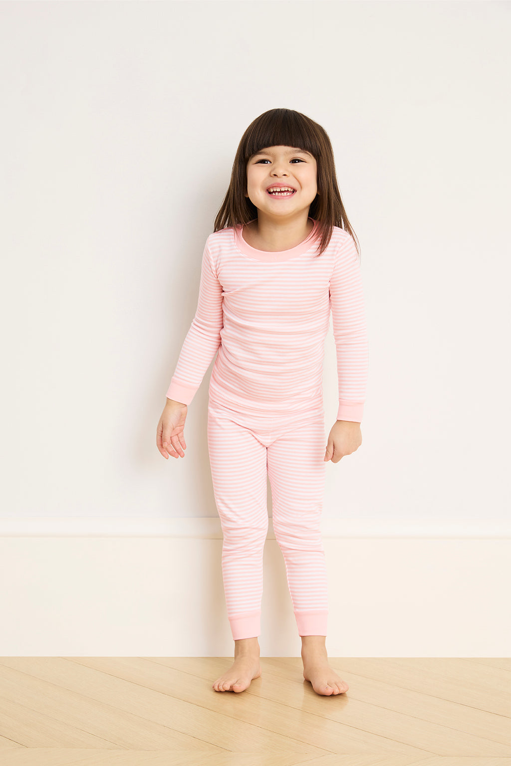 A young girl smiles happily while standing on a wooden floor against a plain light wall, wearing the Kids Long-Long Set in English Rose Stripe—soft Pima cotton pajamas with an elastic waistband.
