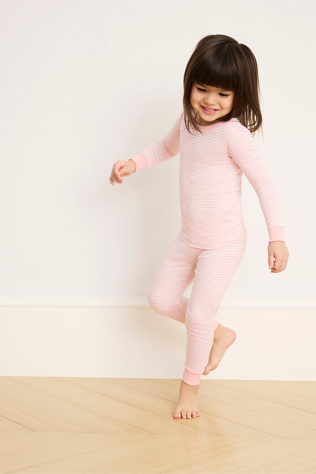 A young child smiles playfully, wearing the Kids Long-Long Set in English Rose Stripe—pink and white striped pajamas made from soft, breathable Pima cotton—against a light wall and wooden floor.