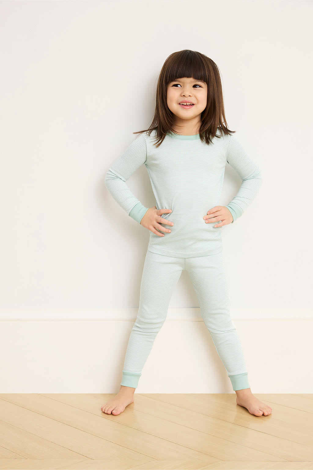 A young child stands on a wooden floor, smiling with hands on hips, wearing the Kids Long-Long Set in Parisian Green against a plain light-colored wall.