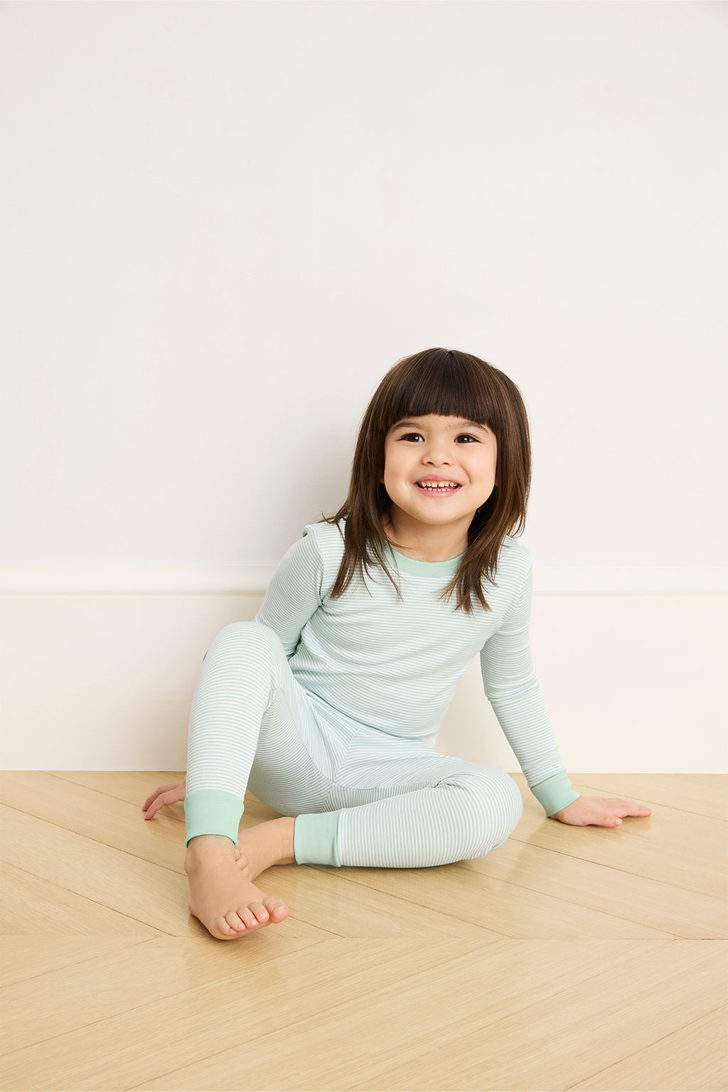 A young child in the Kids Long-Long Set in Parisian Green, crafted from soft Pima cotton, sits on a wooden floor against a white wall, smiling and glancing to the side.