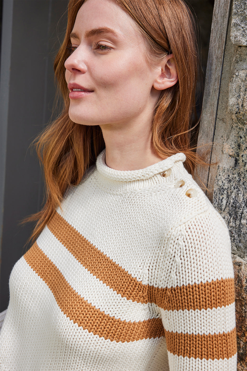 A woman with long red hair, standing outdoors near a stone wall and smiling slightly, wears the Jane Cotton Sweater in Cognac Double Stripe—a chunky cream cotton knit with button details on the shoulder.