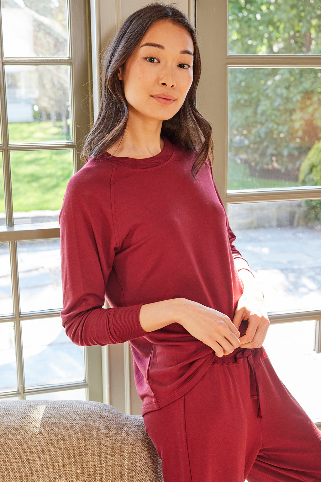 A woman stands indoors by a window, smiling softly as sunlight streams in. She wears the Relax Pants Set in Burgundy, a matching long-sleeve top and pants, gently holding the hem of her shirt.