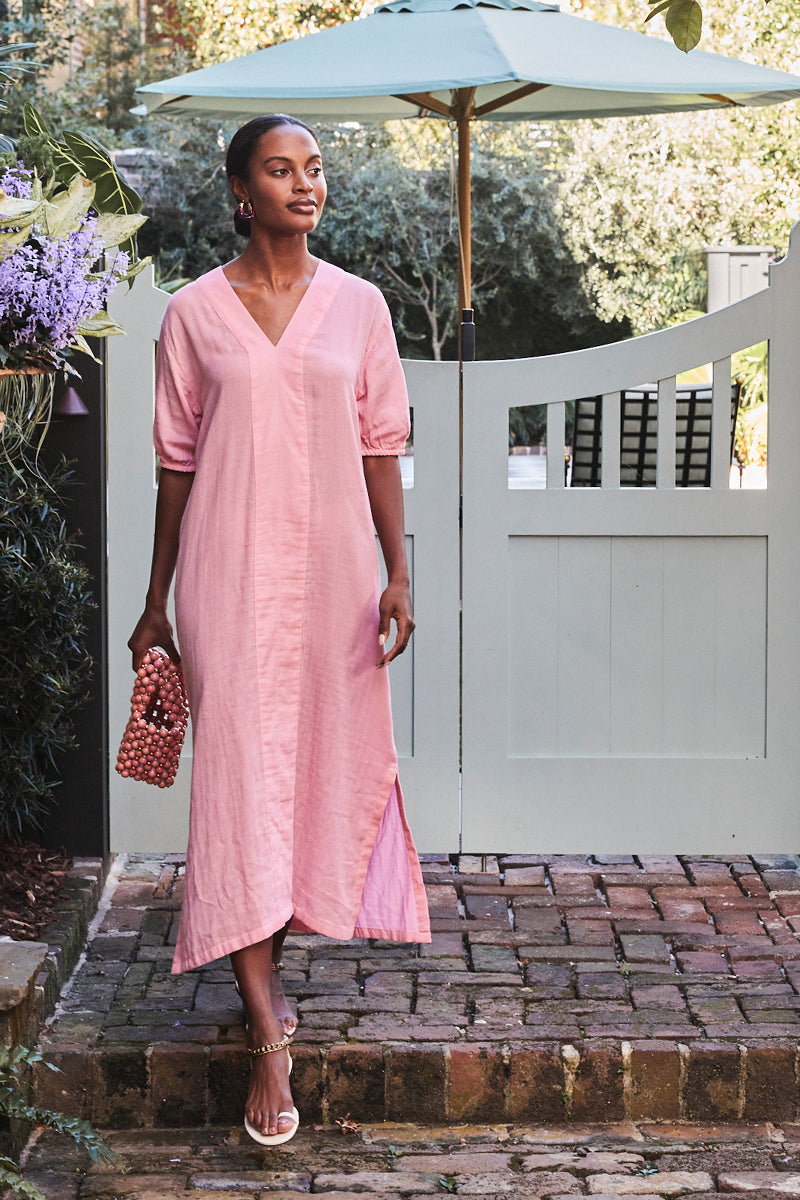 A woman in a coral Gauze Caftan and sandals stands on a brick path, holding a beaded bag near a pale green gate and umbrella, surrounded by greenery.