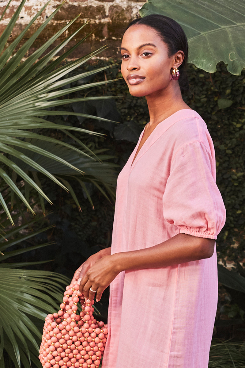 A woman stands outdoors amid greenery, wearing the Gauze Caftan in Coral with puffed sleeves. She carries a pink beaded handbag, accessorized with pink hoop earrings, and gazes calmly at the camera.