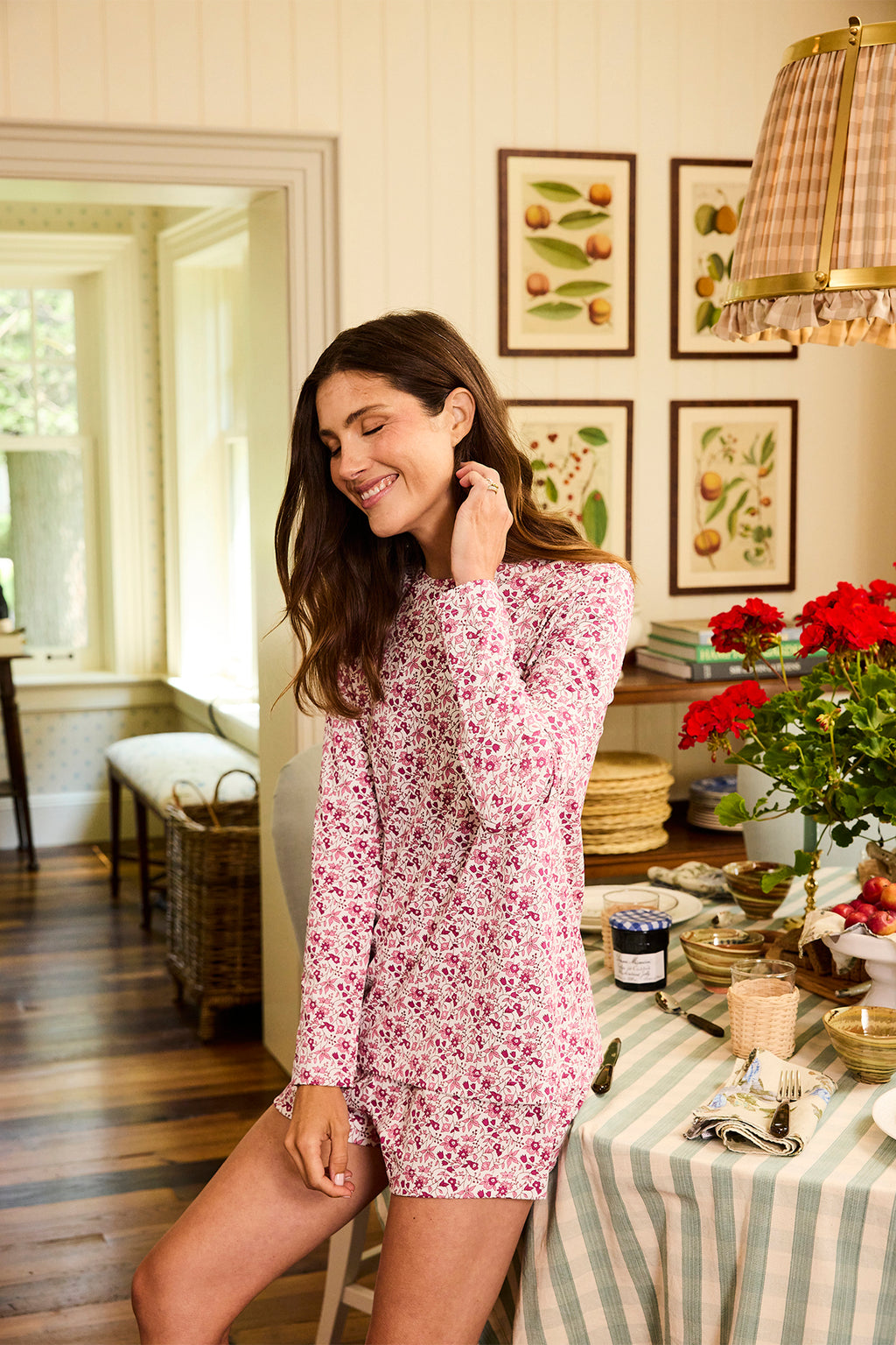 A smiling woman in a Pima Crew Long-Short Set in Dahlia Garden Floral stands in a cozy, sunlit kitchen with checkered curtains, botanical prints, and a table set with food, potted red flowers, and a striped tablecloth.
