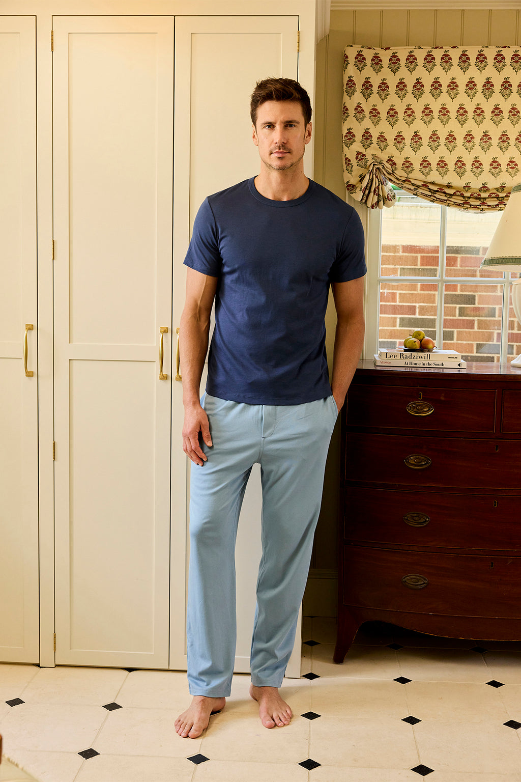 A man stands in the kitchen by a cream cabinet and dark wood dresser, wearing Mens Pima Sleep Pants in Anchor Blue, near a window with a patterned curtain.