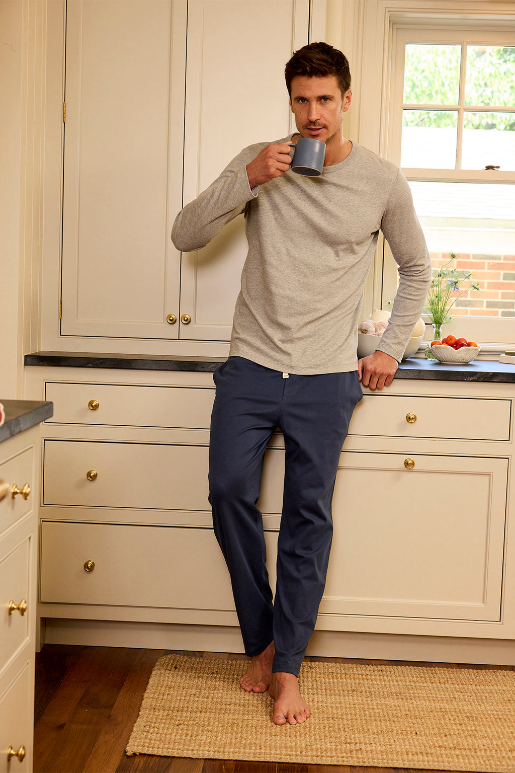 A barefoot man in a light long-sleeve shirt and Mens Pima Sleep Pants in Navy stands in the kitchen, leaning against the counter and drinking from a gray mug as sunlight streams through the window behind him.