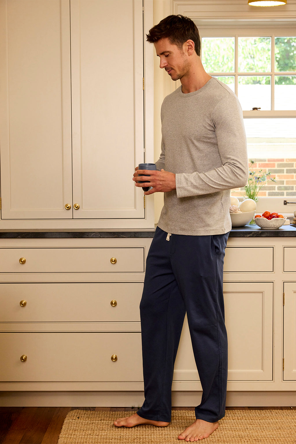 A man wearing a gray long-sleeve shirt and Mens Pima Sleep Pants in Navy stands in a kitchen, holding a mug and looking down as natural light streams through the window behind him.