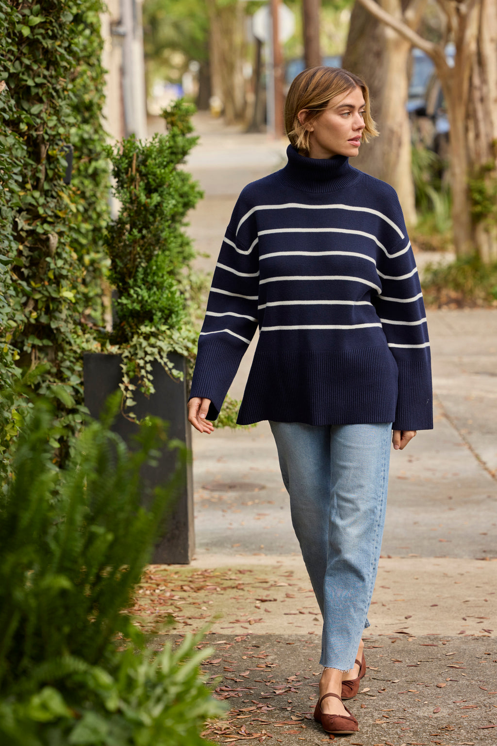 A woman walks outdoors among greenery, wearing the oversized Madeline Sweater in English Navy Banker Stripe, light blue jeans, and brown flats.