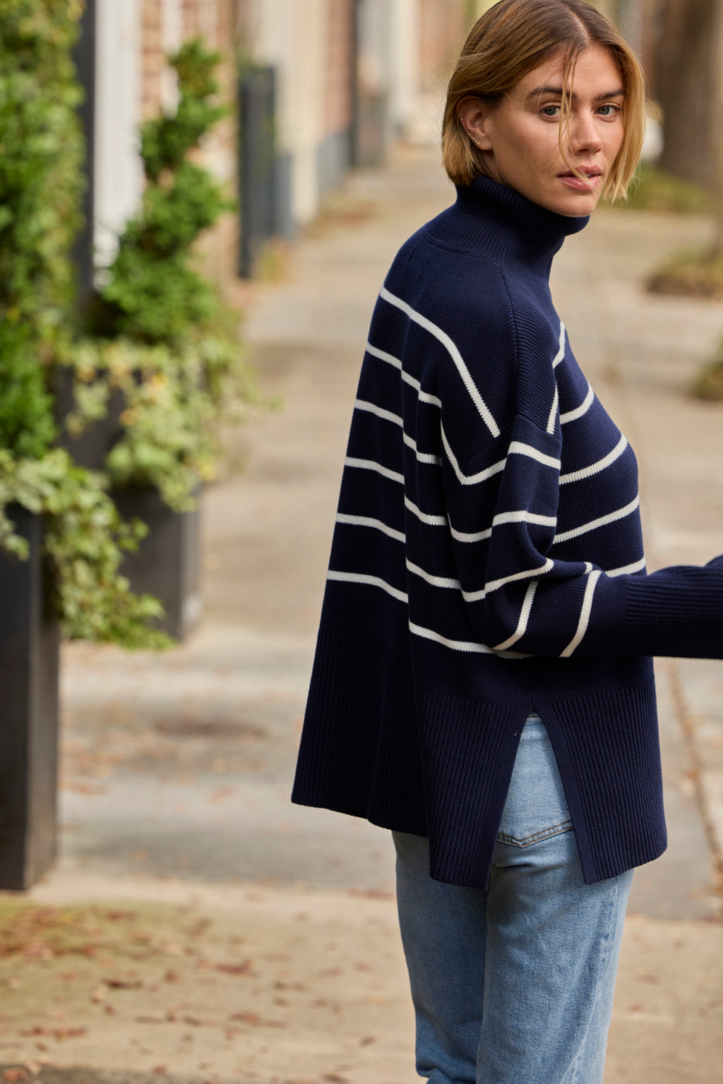 A woman in the Madeline Sweater in English Navy Banker Stripe and light blue jeans stands on a sidewalk, glancing over her shoulder with green plants in the background.