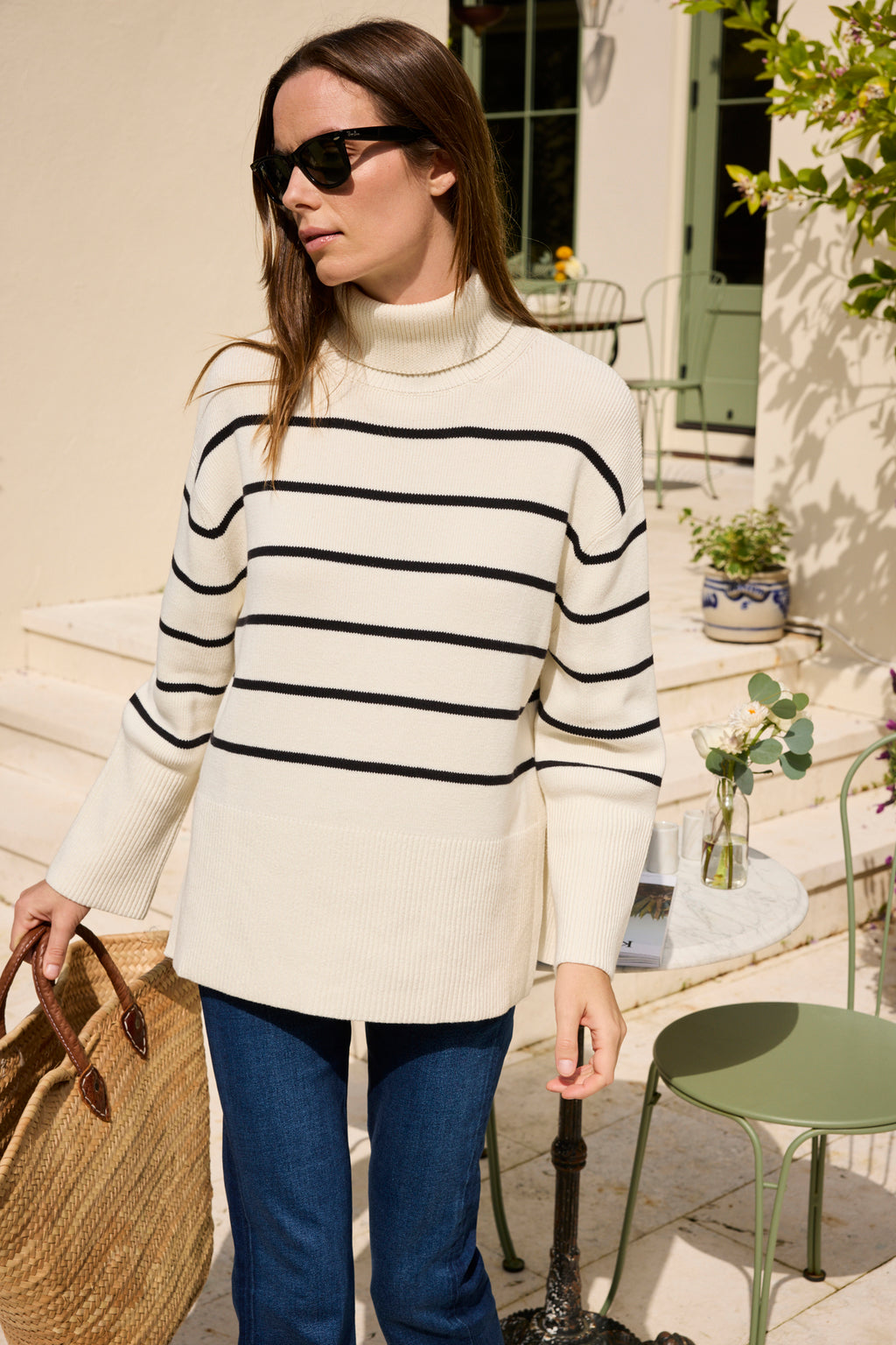 A woman wearing sunglasses and blue jeans stands outdoors by a round table with a vase of flowers, holding a woven tote bag and wearing the Madeline Sweater in Ivory Black Stripe.