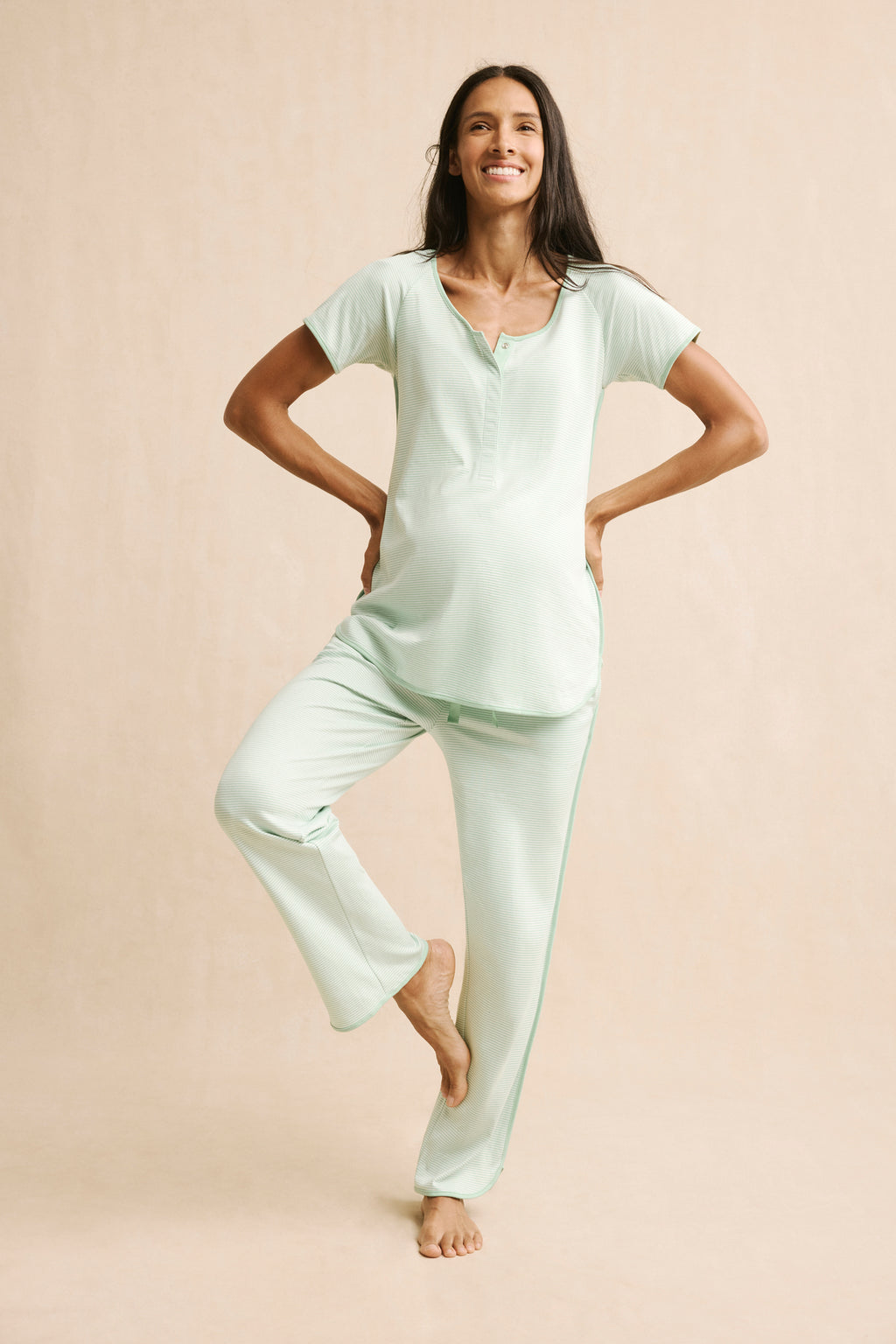 A smiling person stands on one leg in a relaxed yoga pose, wearing the Pima Maternity Short-Long Set in Parisian Green, featuring a matching short-sleeve top, set against a beige background.