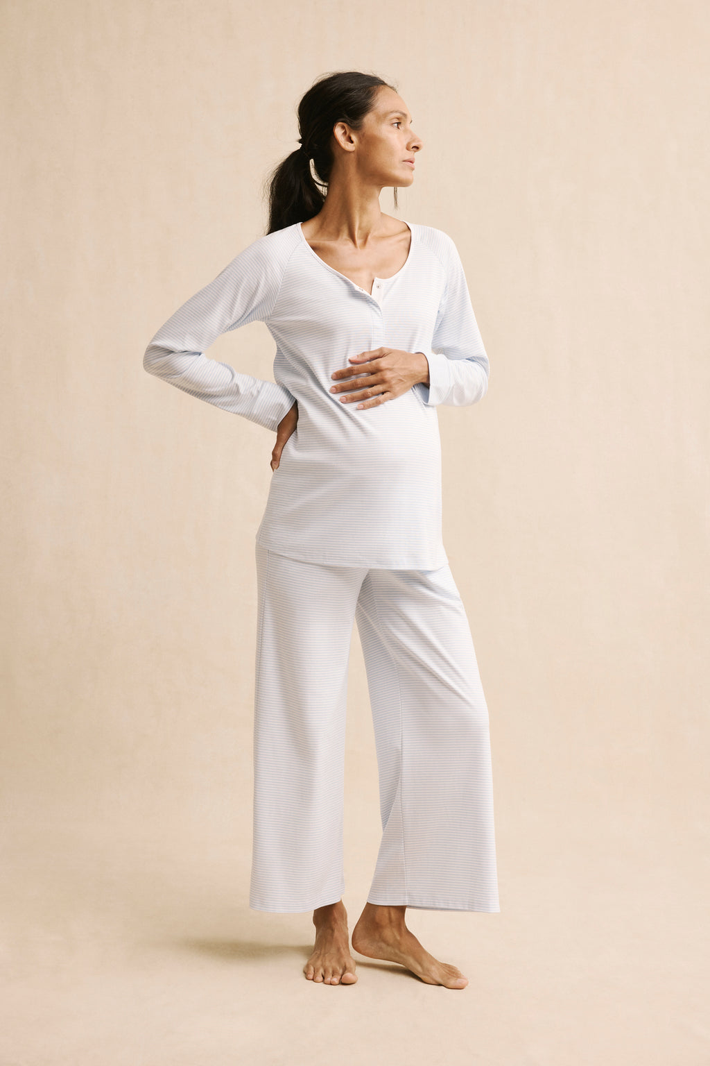 A woman in the Pima Maternity Wide Leg Pajama Set in French Blue, made of soft cotton, stands against a beige background, one hand on her lower back and the other on her belly, looking thoughtfully to the side.