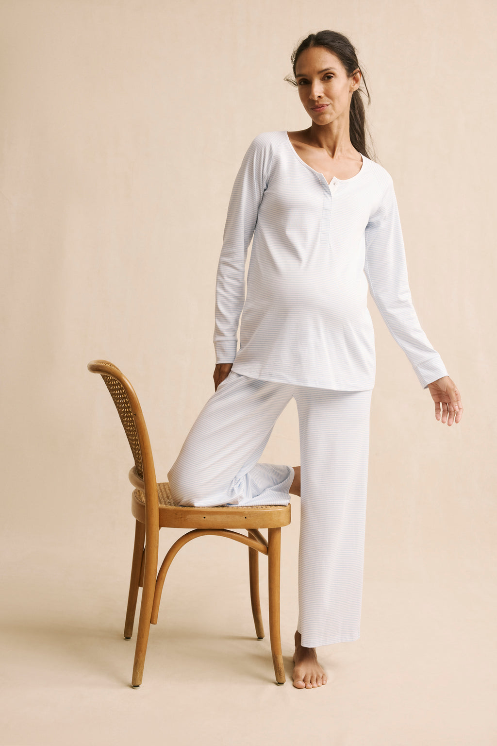 A pregnant woman wears the Pima Maternity Wide Leg Pajama Set in French Blue, standing with one knee on a wooden chair against a neutral beige background.