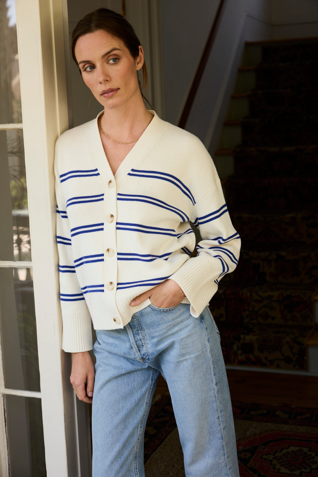 A woman wearing the Nora Cardigan in Ivory Caspian Stripe and light blue jeans stands indoors by a doorway, one hand in her pocket, gazing thoughtfully to the side, with patterned carpeted stairs visible behind her.