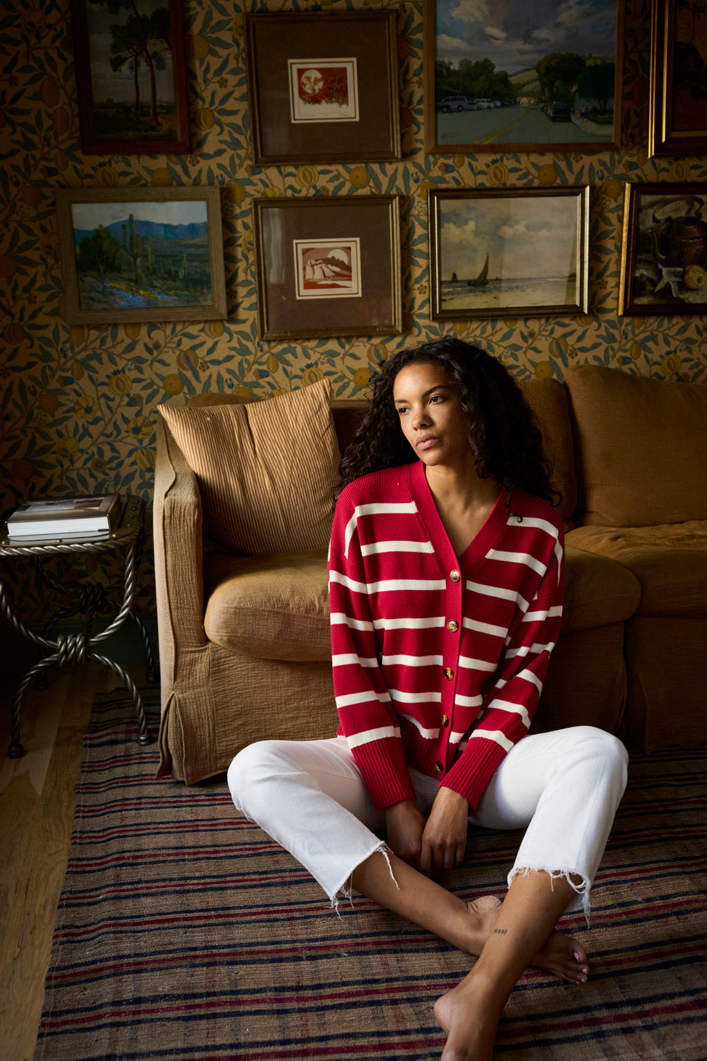 A woman sits on a striped rug in front of a brown sofa, wearing the Nora Cardigan in Ivory Crimson Inverse Stripe and white jeans. Framed art decorates the wallpapered wall behind her.