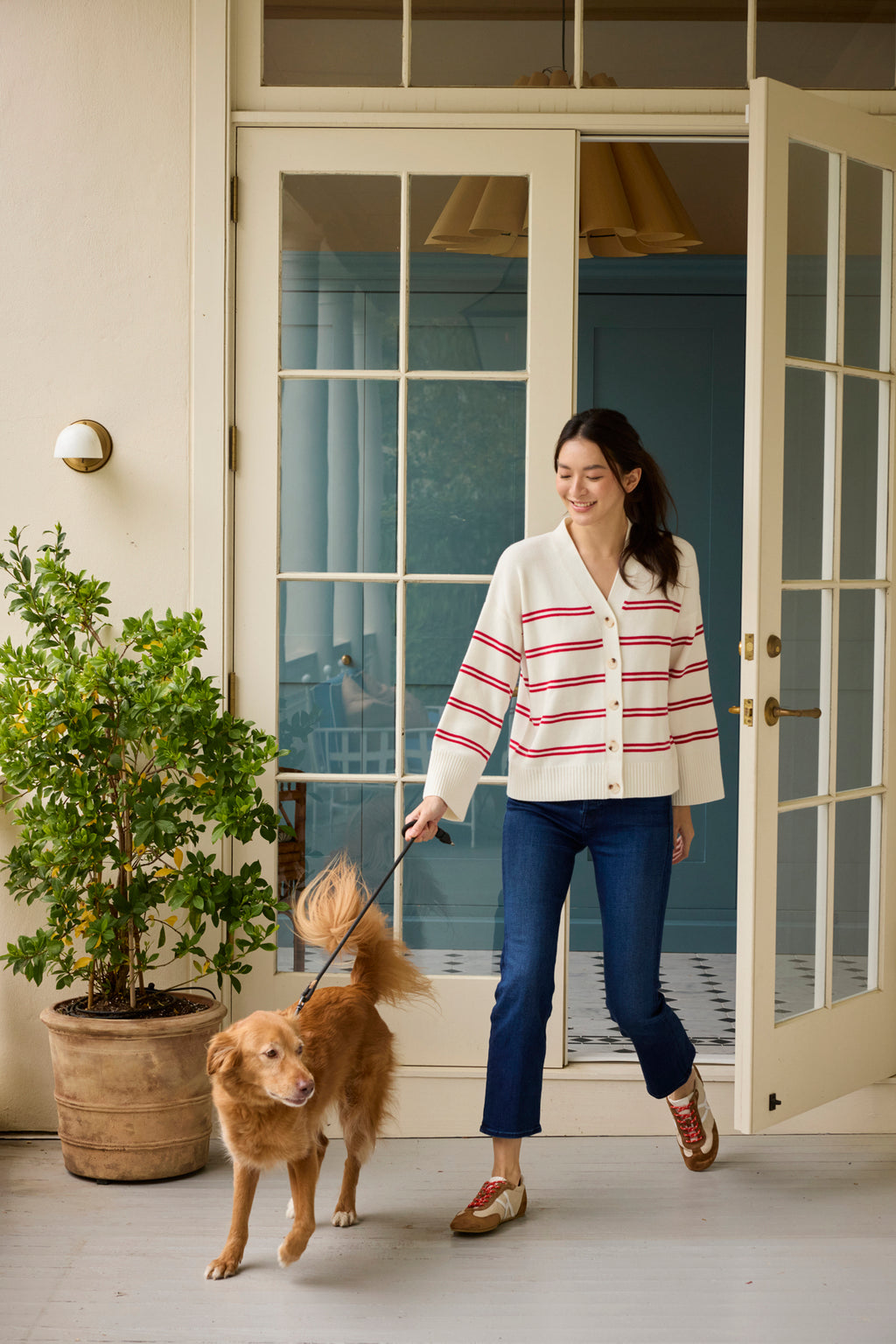 A woman in the Nora Cotton Cardigan in Ivory Red Stripe and blue jeans smiles while walking a golden retriever on a leash outside a house with glass doors and a potted plant nearby.