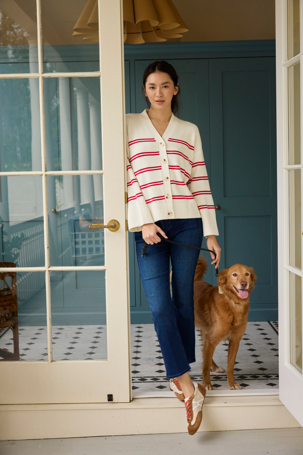 Wearing the Nora Cotton Cardigan in Ivory Red Stripe and blue jeans, a woman stands in a doorway holding her dog