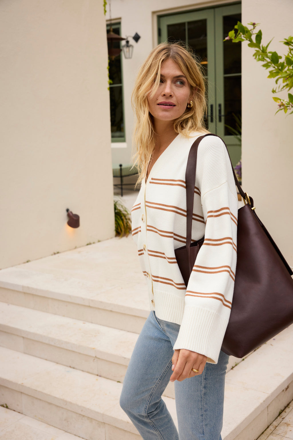 A woman in the Nora Cardigan in Oak Varsity Stripe, light blue jeans, and a large dark brown shoulder bag stands outdoors by white steps and pale green doors.