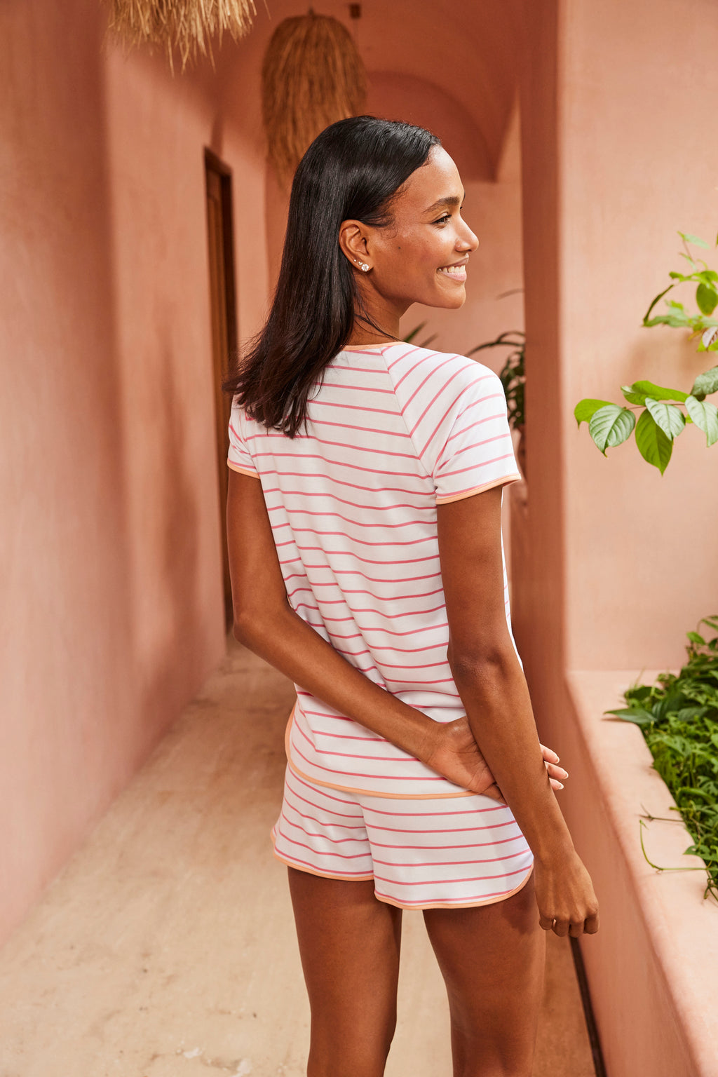 A woman stands in a pink hallway, smiling as she faces away, wearing the Pima Shorts Set in Sunkissed Coral Club Stripe with one arm bent behind her back. Green plants grow along a ledge on the right side.