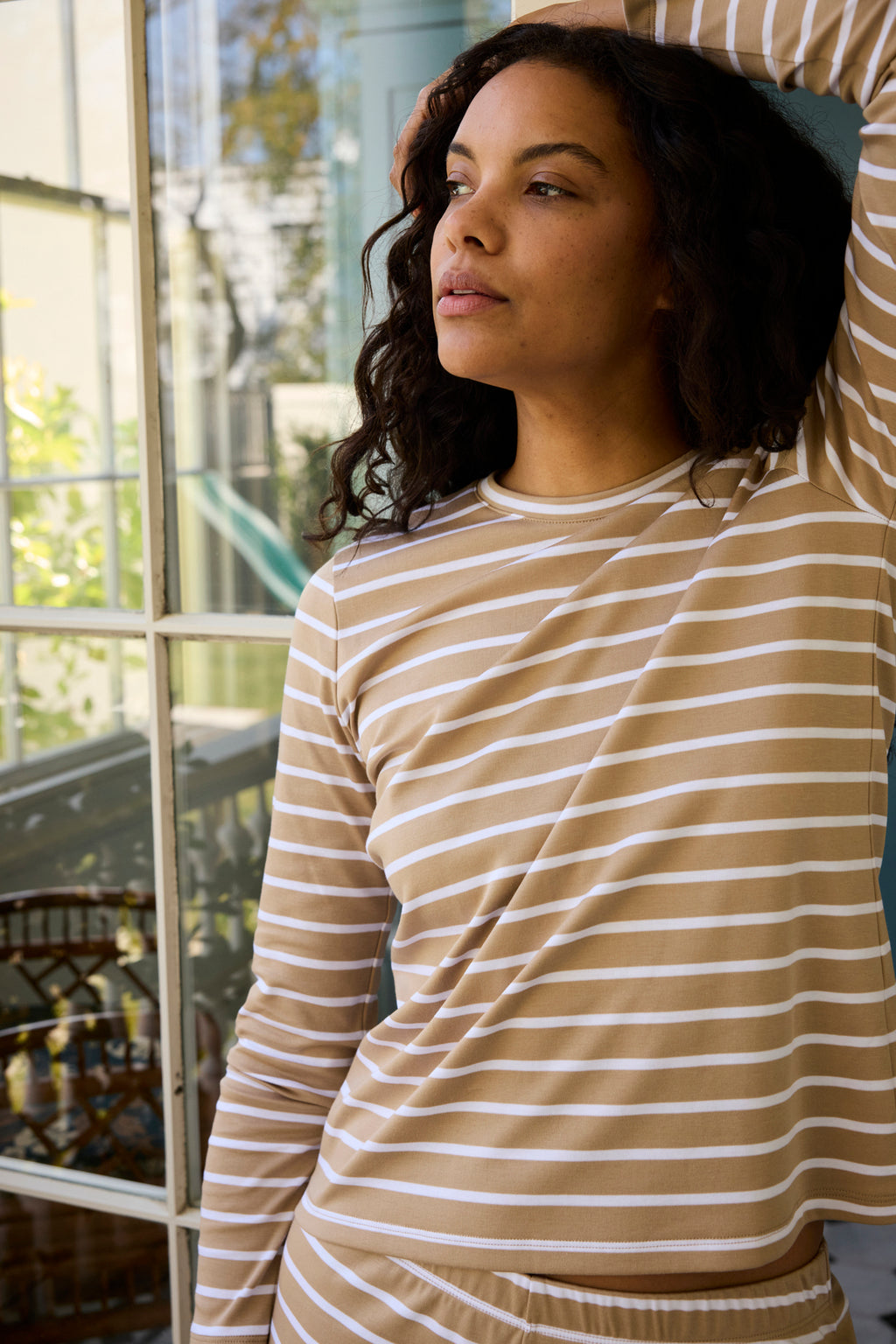 A woman wearing the Pima Crew Long-Short Set in Sandstone Anchor Stripe stands by a window indoors, gazing thoughtfully outside as sunlight streams in, illuminating the greenery beyond.