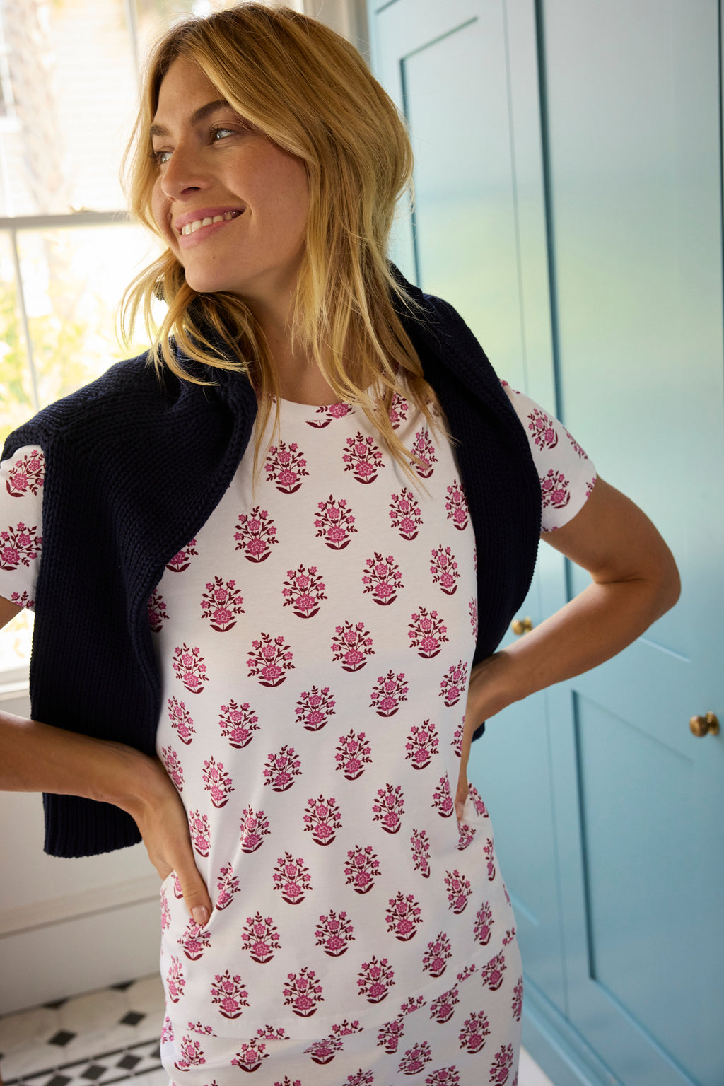 A woman smiles in a room with light blue cabinets, wearing the Pima Crew Short-Long Set in Cabernet Cottage Floral and a dark sweater draped over her shoulders.